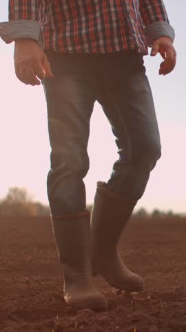 Farmer wearing rubber boots and plaid shirt walking slowly across freshly tilled soil in an open agricultural field during a calm evening on the countryside