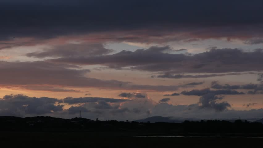 The sky at dusk displays a dramatic palette of colors, with dark, ominous clouds contrasting with streaks of fiery orange and pink. Silhouetted hills and a few distant cars add context to this atmospheric, moody landscape.