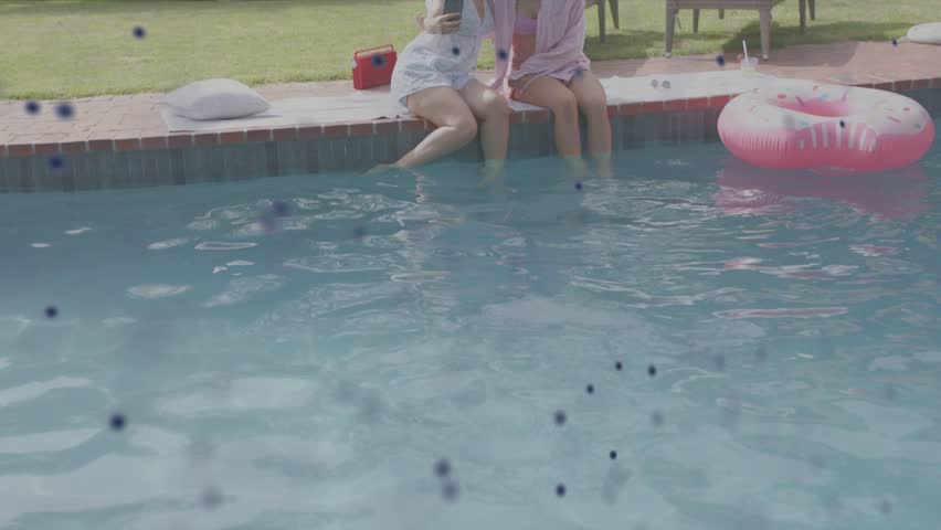 Two women raising phone, leaning pool edge, making social selfie with overlay sweeping across frame. Backyard, outdoor, friends, bonding, sunny, summer, lounge