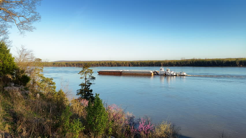 Wide tracking shot of a barge traveling North up the Ohio River near Cave In Rock, IL. The Kentucky River bank is seen across the river.
