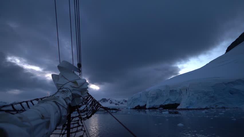 Cinematic POV from the bowsprit of the Schooner Amazone sailing toward massive glaciers in Paradise Bay under a moody twilight sky in Antarctica
