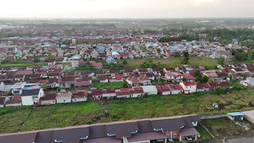 A drone shot capturing a densely populated neighborhood in Indonesia at a misty dawn. The morning mist shrouds the vast expanse of colorful rooftops and the suburban landscape beneath a pale sky.