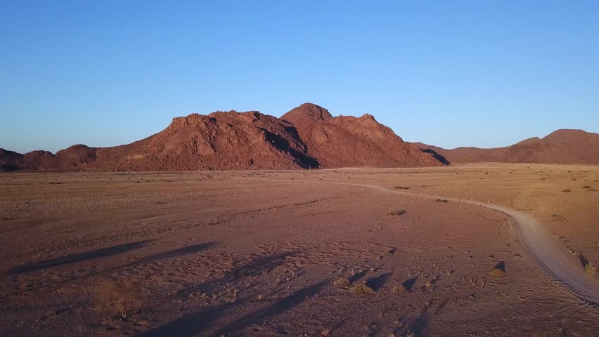 A drone captures the red sand dunes of the Namib Desert. The wide landscape shows the sandy hills under the clear sky. The colors contrast with the dry ground and few plants.