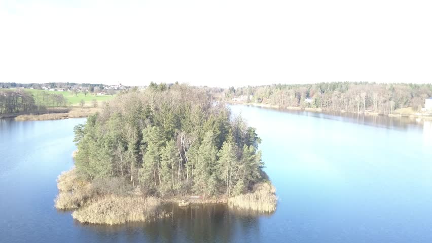 Drone view approaching the lakeshore with trees along the water, natural landscape with calm surface and soft reflections, peaceful atmosphere