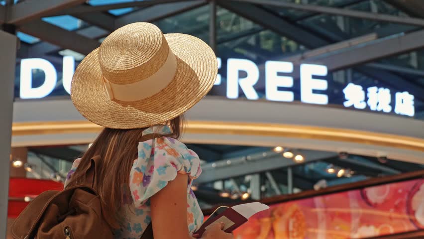 Traveler wearing straw hat holding passport and ticket stands before duty free entrance at airport terminal, international tourism and shopping background for holiday travel advertisement, slow motion