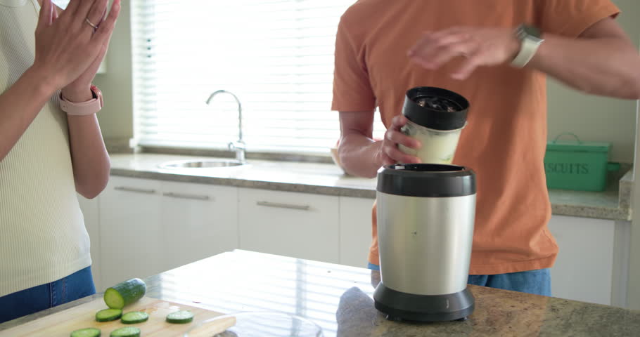 Couple preparing cucumber smoothie on kitchen island, man securing blender while woman placing cups. Healthy, green, juice, glasses, home, daylight, teamwork