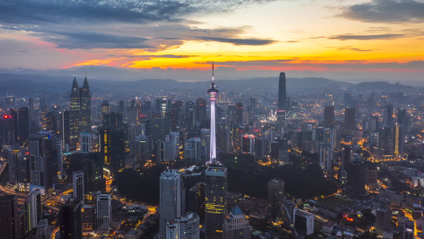Cinematic aerial tilt-up hyperlapse of a modern metropolitan skyline at dawn in Kuala Lumpur, Malaysia. Features a dramatic sunrise with strong golden light and visible sun rays piercing through the morning haze. The urban landscape includes glass skyscrapers and busy roads as the city wakes up. High-quality ProRes footage ideal for business, finance, and travel concepts.