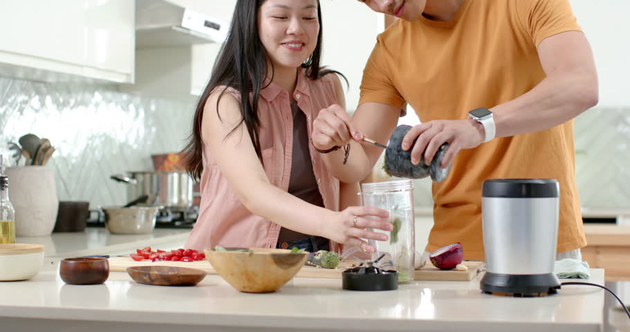 Asian couple making smoothie at kitchen island, man grabbing jar scooping berries into blender cup. Fruit, collaboration, domestic, bright, lifestyle, countertop, healthy