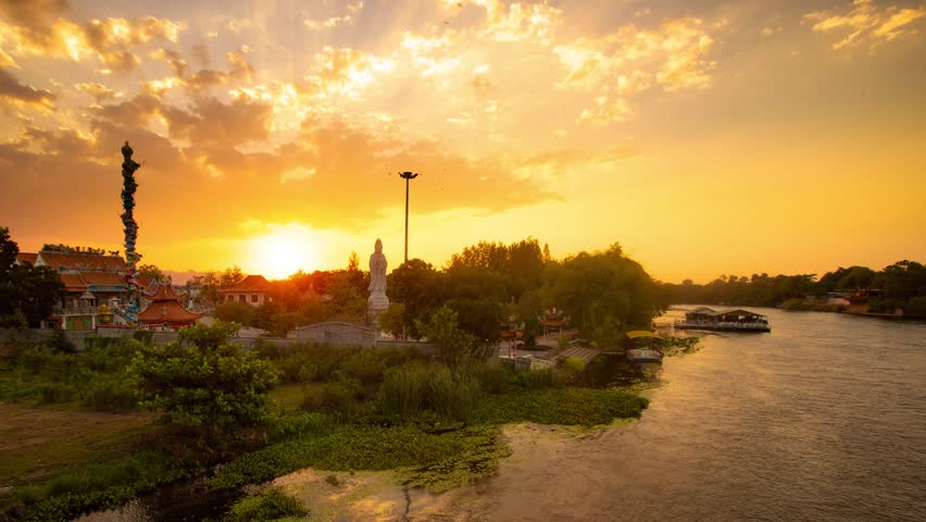 Timelapse of sunset over Guanyin Fushou Temple and River Kwai in Kanchanaburi Thailand. View from railway bridge with reflections, warm light and peaceful atmosphere.