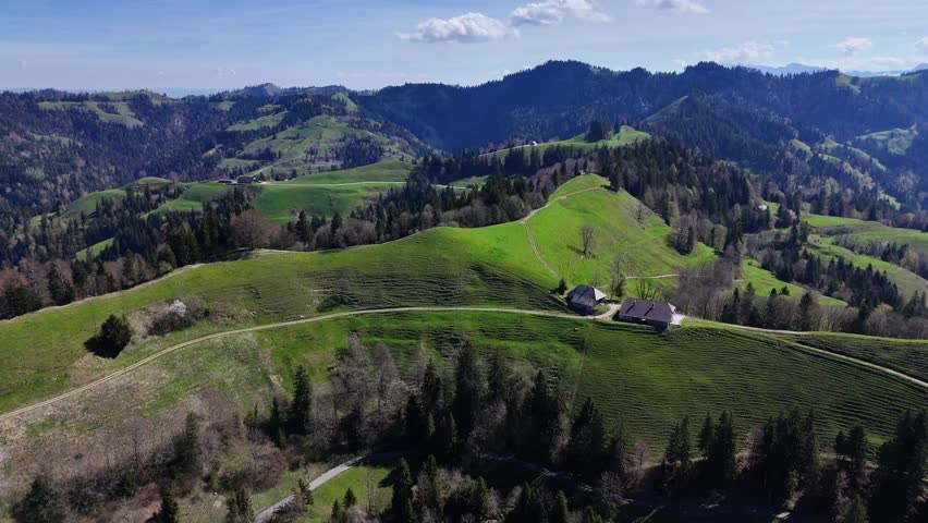 Panoramic view of green rolling hills, trees, and farms in Emmental, Switzerland on a bright spring day with dramatic clouds and distant ridgelines.
