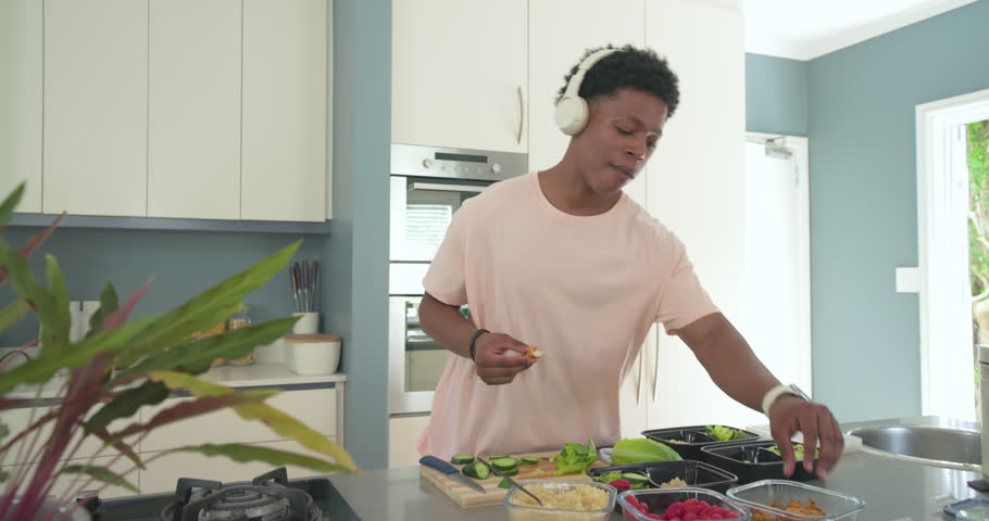 African American man 20s tasting and filling meal boxes at kitchen island with headphones for lunch. Prep, containers, portions, cucumber, greens, raspberries, home