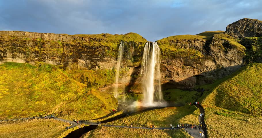Aerial view of Seljalandsfoss in South Iceland, with visitor footpaths, glowing mossy cliffs, and the famous waterfall plunging in rich evening light.