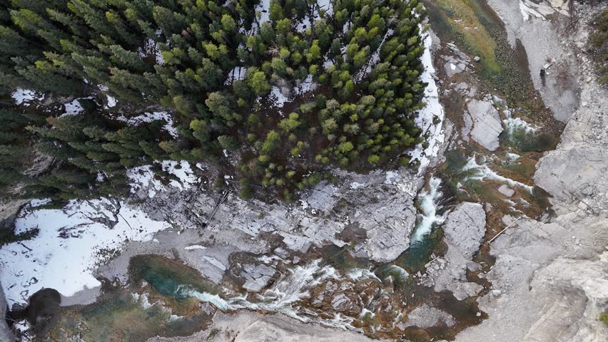 A cinematic collection of aerial drone images capturing the rugged beauty of the Rocky Mountains during the transition from winter to early spring. From a top-down perspective, a winding alpine river cuts through a rocky canyon, its turquoise and emerald waters flowing over layered stone, forming small rapids, cascades, and calm pools. The clarity of the water reveals intricate textures beneath the surface, while braided channels, gravel bars, and exposed riverbeds create organic patterns that resemble natural abstract art.

Dense evergreen forests surround the river, forming deep green clusters that contrast with pale rock, sand, and lingering snow. From above, the trees appear textured and layered, following the contours of the terrain and emphasizing elevation changes across the landscape. Patches of snow and thin ice remain along shaded banks and forest edges, highlighting the seasonal shift and adding visual contrast between frozen and flowing elements.

The aerial perspective reveals dramatic geological features, including exposed rock slabs, eroded canyon walls, and natural river bends that wrap around forested islands and peninsulas. These formations shape the movement of the water while adding depth and scale to the composition. Soft, diffused lighting enhances natural colors and textures, while distant mountain ridges and cloud-filled skies introduce a sense of atmosphere and vastness.

These high-resolution drone images showcase untouched wilderness with no visible human presence, evoking a sense of isolation, tranquility, and raw natural beauty. Ideal for themes such as outdoor adventure, environmental conservation, travel, and landscape design, the collection highlights the Rocky Mountains through a unique aerial perspective, capturing patterns, textures, and seasonal transitions that are rarely seen from ground level.
