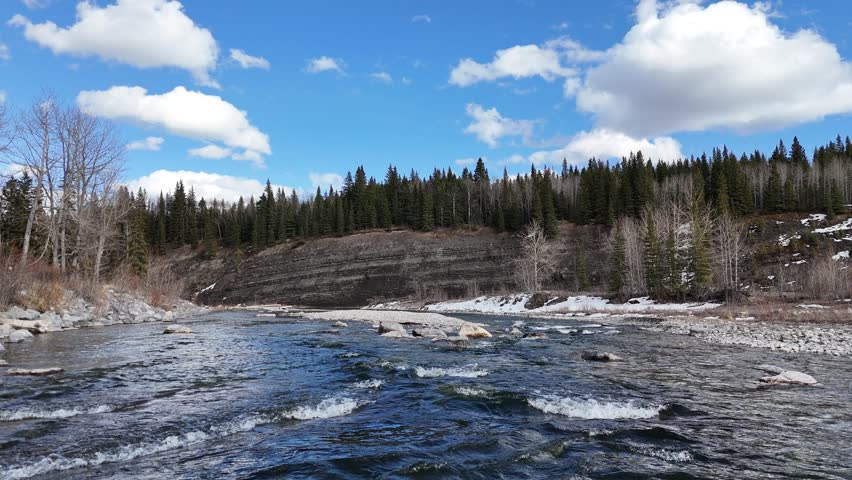 A cinematic collection of aerial drone images capturing the rugged beauty of the Rocky Mountains during the transition from winter to early spring. From a top-down perspective, a winding alpine river cuts through a rocky canyon, its turquoise and emerald waters flowing over layered stone, forming small rapids, cascades, and calm pools. The clarity of the water reveals intricate textures beneath the surface, while braided channels, gravel bars, and exposed riverbeds create organic patterns that resemble natural abstract art.

Dense evergreen forests surround the river, forming deep green clusters that contrast with pale rock, sand, and lingering snow. From above, the trees appear textured and layered, following the contours of the terrain and emphasizing elevation changes across the landscape. Patches of snow and thin ice remain along shaded banks and forest edges, highlighting the seasonal shift and adding visual contrast between frozen and flowing elements.

The aerial perspective reveals dramatic geological features, including exposed rock slabs, eroded canyon walls, and natural river bends that wrap around forested islands and peninsulas. These formations shape the movement of the water while adding depth and scale to the composition. Soft, diffused lighting enhances natural colors and textures, while distant mountain ridges and cloud-filled skies introduce a sense of atmosphere and vastness.

These high-resolution drone images showcase untouched wilderness with no visible human presence, evoking a sense of isolation, tranquility, and raw natural beauty. Ideal for themes such as outdoor adventure, environmental conservation, travel, and landscape design, the collection highlights the Rocky Mountains through a unique aerial perspective, capturing patterns, textures, and seasonal transitions that are rarely seen from ground level.