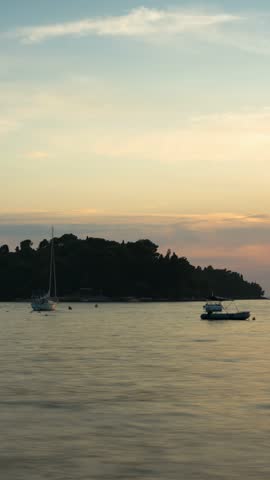 Sunset Over the Adriatic Sea in Rovinj, Croatia, Scenic Coastal Landscape with Calm Water, Silhouetted Shoreline, and Stone Pier at Dusk Rovinj, Croatia - August 16, 2025: A serene wide-angle sequence capturing the golden hour in Rovinj, Croatia. The scene transitions across the harbor, showing sailboats anchored near lush Mediterranean greenery and a traditional stone pier in the foreground. Warm sunset light fills the sky with hues of orange and gold, reflecting on the rippling sea. Perfect for travel documentaries, tourism marketing, and cinematic b-roll.