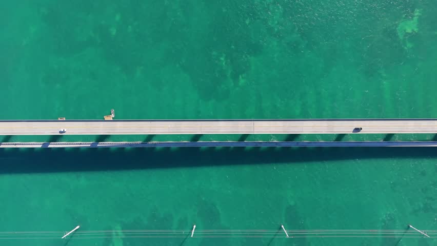 A breathtaking aerial drone perspective of the iconic Seven Mile Bridge in the Florida Keys. The footage captures the parallel stretch of the modern Overseas Highway and the historic Old Seven Mile Bridge as they cross the crystal-clear emerald and turquoise waters of the Atlantic Ocean. This high-angle view highlights the incredible engineering scale and the stunning tropical landscape connecting the Middle and Lower Keys. Perfect for travel documentaries, automotive marketing, and Florida tourism showcases.