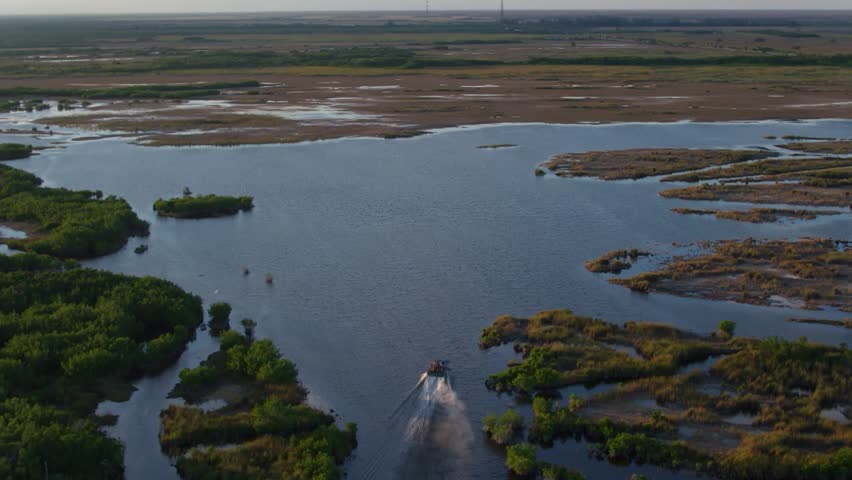 A dynamic high-angle aerial view capturing a classic Florida airboat as it speeds through the pristine wilderness of the Everglades. The footage shows the airboat creating a white water wake as it glides over the shallow sawgrass marshes and through narrow freshwater channels. Surrounded by the vast, untouched tropical wetlands and a clear blue horizon, this shot perfectly illustrates the adventure and scale of the Florida swamplands. Ideal for travel documentaries, adventure tourism promos, and nature films focused on the unique American Everglades ecosystem.