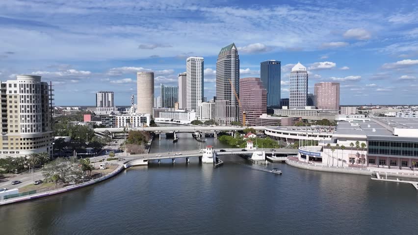 A stunning high-angle aerial view of the vibrant downtown Tampa skyline in Florida. The footage captures the high-rise architecture of the business district as it towers over the shimmering waters of the Hillsborough River. Prominently featured is the Madame Fortune Taylor Bridge (historically known as the Laurel Street Bridge), which connects the urban core to the lush Julian B. Lane Riverfront Park. This professional establishing shot highlights the scenic Riverwalk area and the blend of modern urban living with coastal Florida beauty. Ideal for travel documentaries, real estate marketing, and corporate video projects.