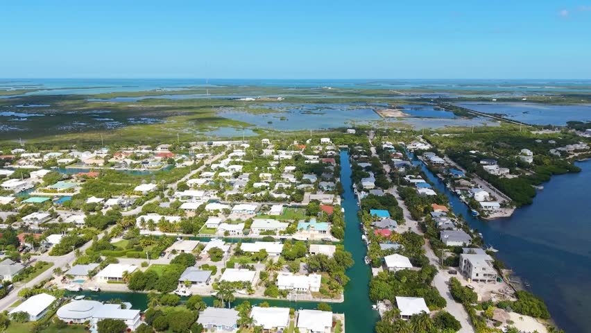 A stunning high-angle aerial view of a typical residential neighborhood in the Florida Keys. The footage showcases the unique urban planning of the archipelago, featuring private homes situated along deep-water man-made canals that provide residents with direct boating access to the Atlantic Ocean or Gulf of Mexico. Lush tropical greenery, palm trees, and clear turquoise waters define this coastal community. This professional establishing shot is perfect for real estate marketing, travel documentaries, and lifestyle content highlighting island living and maritime culture.