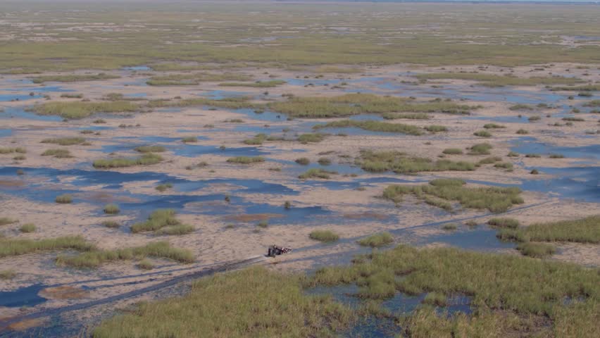 An action-oriented aerial tracking shot of a traditional Florida airboat as it maneuvers through the vast "River of Grass." The footage captures the high-speed movement of the vessel across shallow freshwater marshes, throwing up a distinctive white spray against the vibrant green vegetation. The expansive landscape stretches to the horizon under a clear sky, showcasing the unique beauty and scale of the Everglades ecosystem. This professional-grade footage is perfect for adventure travel promos, environmental documentaries, and high-energy Florida tourism content.