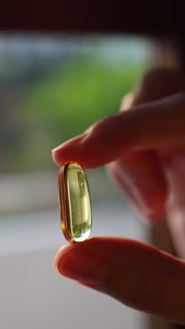 Macro shot of a hand holding a yellow fish oil capsule or vitamin D supplement against a blurred background. Close-up of a golden transparent softgel pill. Health and omega-3 stock footage.