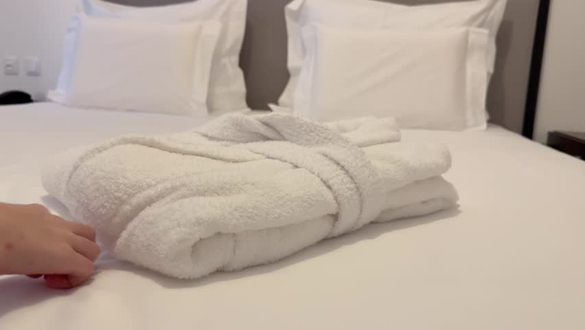 Person removing white bathrobe from neatly made bed, slow motion close up