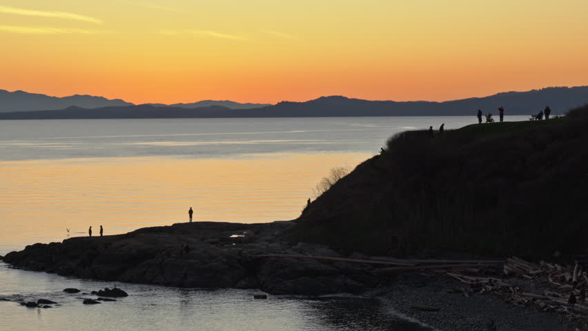 Golden hour sunset over ocean coastline with bright orange sky, silhouetted hill and people walking along the shore and ridge. Scenic coastal landscape with dramatic l ight, calm atmosphere and natural