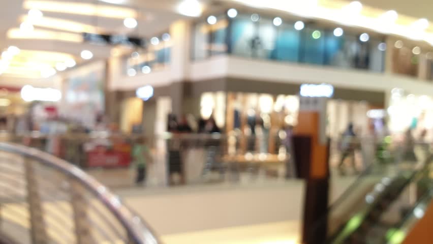 Unrecognizable people walk through a bright defocused shopping center, soft glowing lights and moving silhouettes create a busy atmosphere.