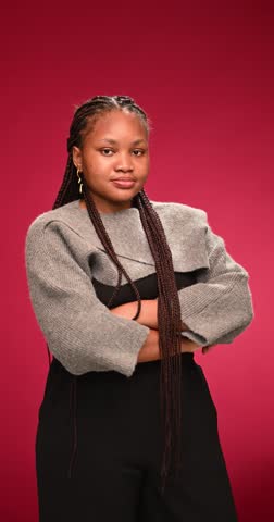 Happy young woman with long braids stands with folded arms against a red background in a lifestyle and fashion portrait highlighting confidence, expression and modern style.