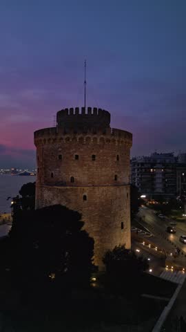 Aerial vertical view of the illuminated White Tower of Thessaloniki at twilight. Beautiful purple sunset sky with an airplane flying in the background.