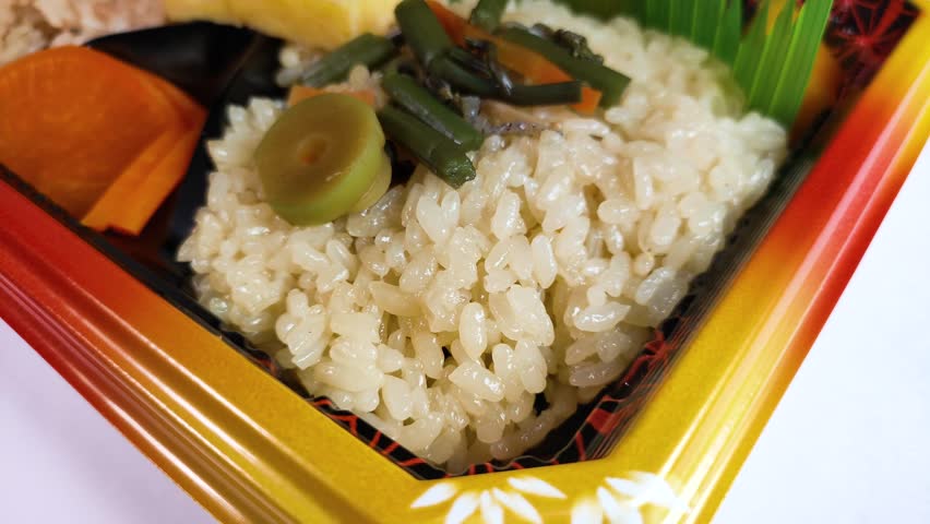 Rotating Japanese bento lunch box with wild mountain vegetable okowa rice and side dishes in a plastic pack on a white background