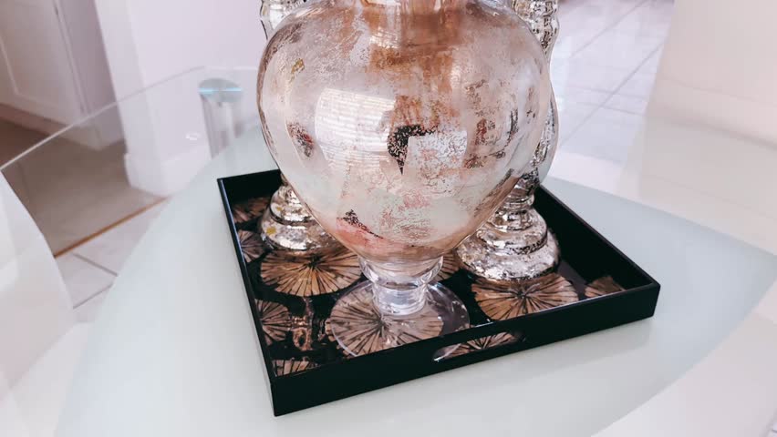 A reflective mercury glass vase holds a full arrangement of dried white baby breath stems, positioned on a clear glass dining table. The metallic surface of the vase features a textured, antiqued finish that softly reflects surrounding light and interior elements. Matching silver candle holders stand nearby, adding height and symmetry to the arrangement. In the background, white cabinetry and neutral flooring create a clean, modern backdrop that emphasizes the soft white florals and shimmering metal surfaces. The dried stems spread outward in a cloud like form, adding delicate texture above the structured base of the vase. The contrast between transparent glass, polished silver tones, and organic dried florals creates a balanced composition centered on refined interior styling and subtle decorative detail.