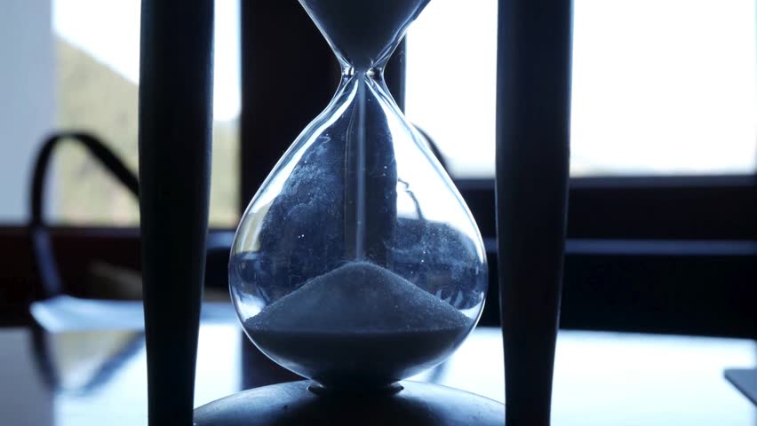 A close-up of an hourglass with sand flowing on a table next to a window with natural light shining through, creating a serene and contemplative atmosphere with dark shadows