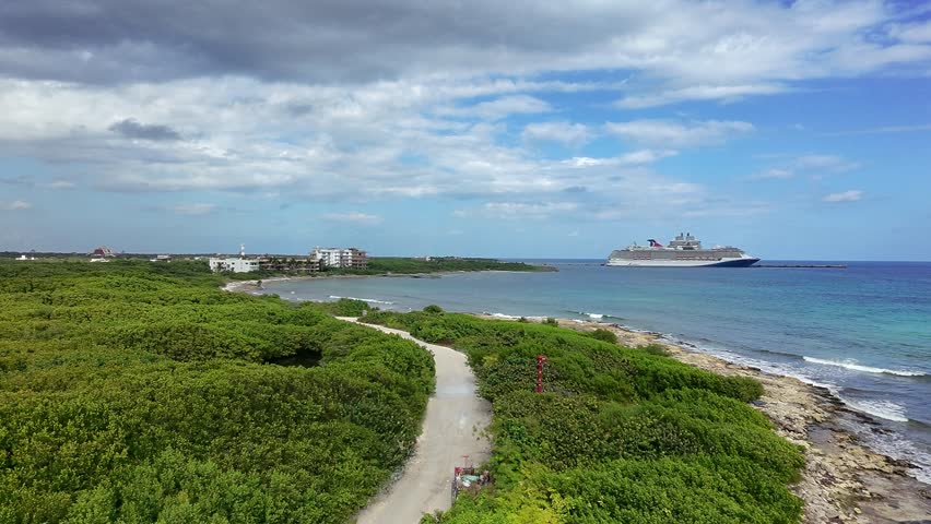 Aerial drone footage capturing the tropical coastline of Mahahual in Costa Maya, Quintana Roo, Mexico, facing north from downtown. The scene features lush coastal vegetation, a winding shoreline road, and clear turquoise Caribbean waters stretching to the horizon. A large cruise ship is docked offshore at the Costa Maya cruise port, one of Mexico’s major Caribbean cruise destinations designed specifically for passenger ships. The area, once a small fishing village, has grown into a popular tourist destination known for its relaxed atmosphere, pristine beaches, and proximity to ancient Mayan sites. This scenic seascape highlights the contrast between natural coastal beauty and modern cruise tourism infrastructure, making it ideal for travel, vacation, and tourism concepts.