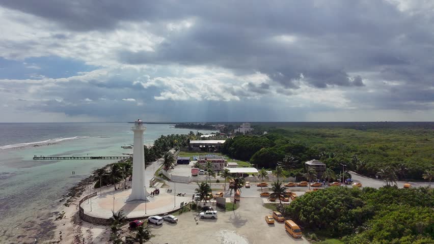 Aerial drone view of the Faro de Mahahual lighthouse located in Mahahual, Quintana Roo, Mexico, along the Caribbean Sea coastline. This white coastal lighthouse, approximately 22 meters tall, is a recognizable landmark of the Costa Maya region and a popular attraction for visitors exploring this once small fishing village turned growing tourist destination.
The footage captures a scenic tropical landscape featuring turquoise waters, palm-lined shores, a pier extending into the sea, and the surrounding coastal village. Sun rays breaking through dramatic clouds add atmosphere and depth, enhancing the natural beauty of this Caribbean setting.
Mahahual is known for its proximity to the Mesoamerican Barrier Reef and cruise port, making it a key destination for tourism in southeastern Mexico.
This video is ideal for themes related to travel, tourism, tropical destinations, coastal environments, vacation lifestyle, and nature. Suitable for commercial use in advertising, promotional content, and editorial storytelling about Mexico’s Caribbean coast.