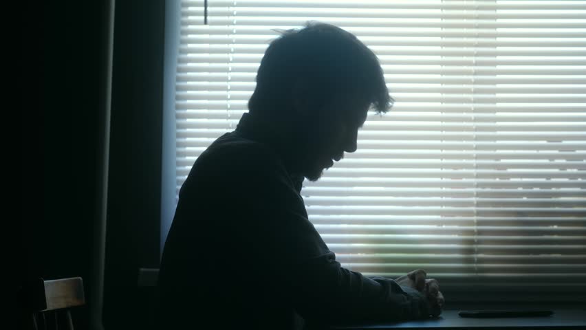 Sad and depressed man in silhouette sits alone at table in dark room, his head in his hands, representing despair, anxiety, loneliness, mental health issues. Depressed, sad man sitting in dark room