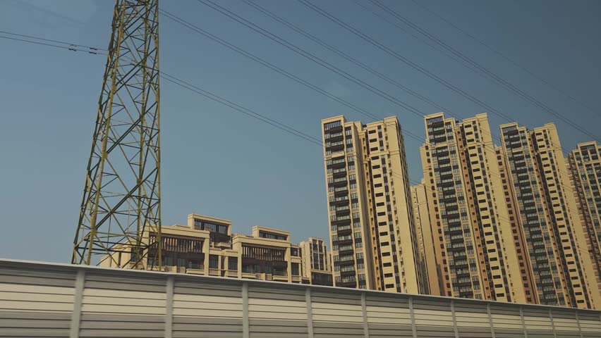 Urban scene showing a series of high-rise buildings next to a sound barrier wall, viewed from a vehicle in motion under a clear blue sky