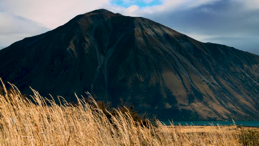 A view of a large dark mountain rising above a serene lake with golden grass in the foreground.