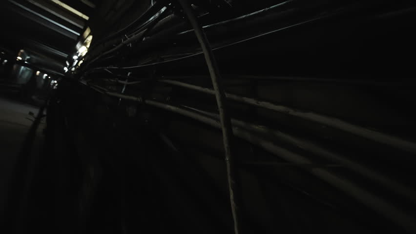 Gritty industrial utility tunnel with dense pipes, cables, and conduits along dark walls, dimly lit by scattered fixtures, evoking an eerie maintenance corridor perspective.