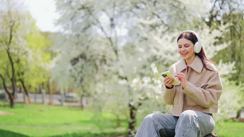 Woman enjoys music with headphones in a beautiful blooming park. Expresses joy, happiness, and serenity amidst nature. Listening to favorite songs creates peaceful outdoor experience.