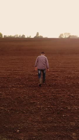 Solitary farmer walking across vast freshly plowed brown field at sunset, contemplating rural landscape and seasonal work beneath expansive overcast sky