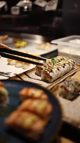 Vertical screen buffet sushi roll being picked with tongs and placed on plate close up self service dining fresh asian food choice minimal motion calm restaurant atmosphere