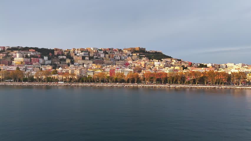 Aerial view of Naples, Italy, with the tree-lined waterfront, calm bay, and stacked hillside neighborhoods catching soft golden evening light.