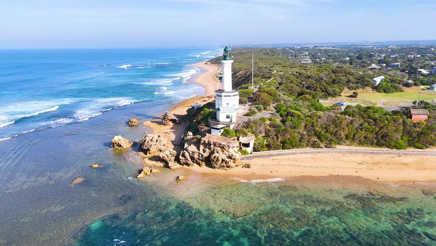 Lighthouse on coastal rocks with ocean waves and a clear sky.