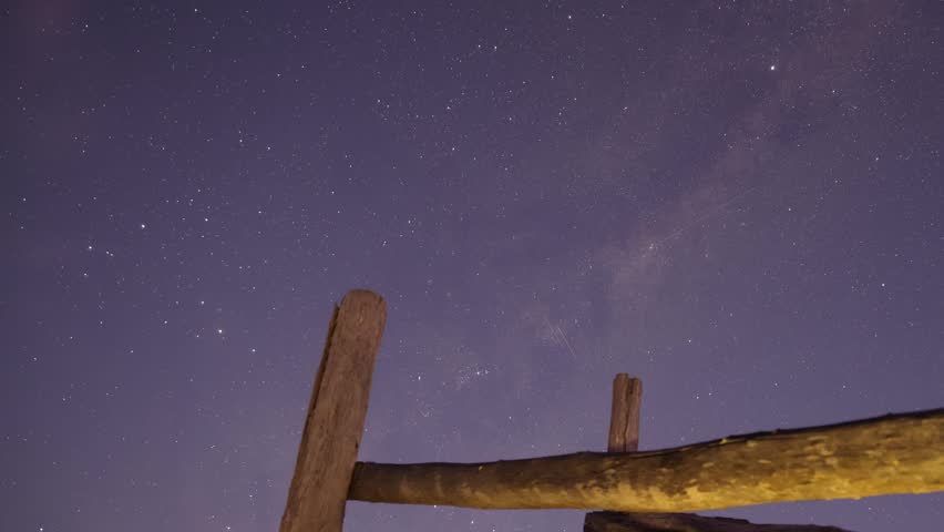 Milky way moving across night sky with stars over rustic fence.