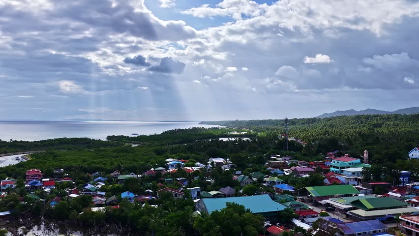 Sunlight breaks through clouds over a vibrant coastal village, highlighting lush greenery and colorful rooftops.