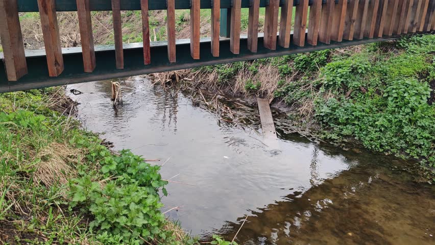 Small stream flowing under wooden pedestrian bridge with metal supports. Rural infrastructure crossing water channel with natural vegetation on banks.