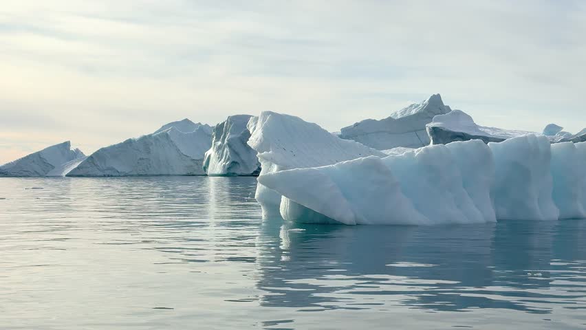 Icebergs drift slowly in the water. The blue ice contrasts with the gray sky. The scene captures the beauty of nature and the movement of the ice on a calm day.