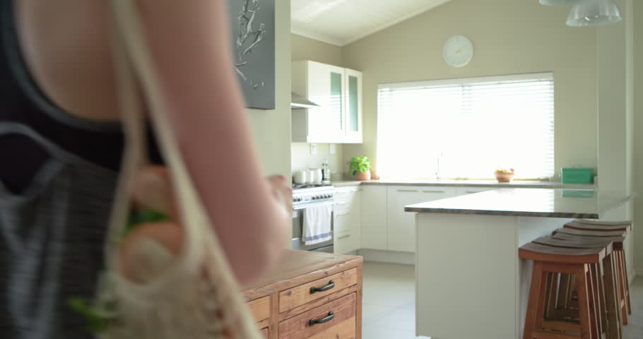 Adult female in athletic wear entering kitchen carrying mesh bag after shopping, unpacking produce. Sunlit, island, stove, barstools, smartwatch, vegetables, fruits