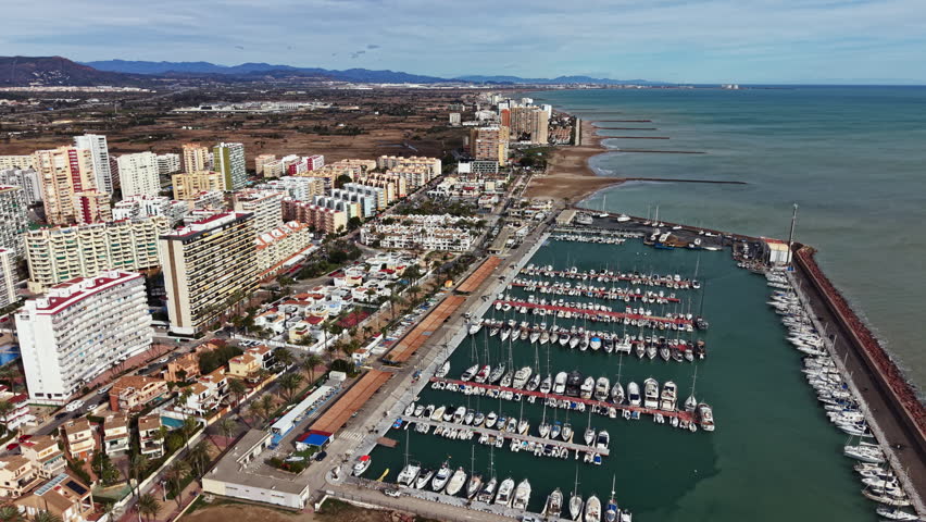 Many boats are moored in the harbor while buildings rise nearby. The sea stretches along the coast and hills are visible in the distance.